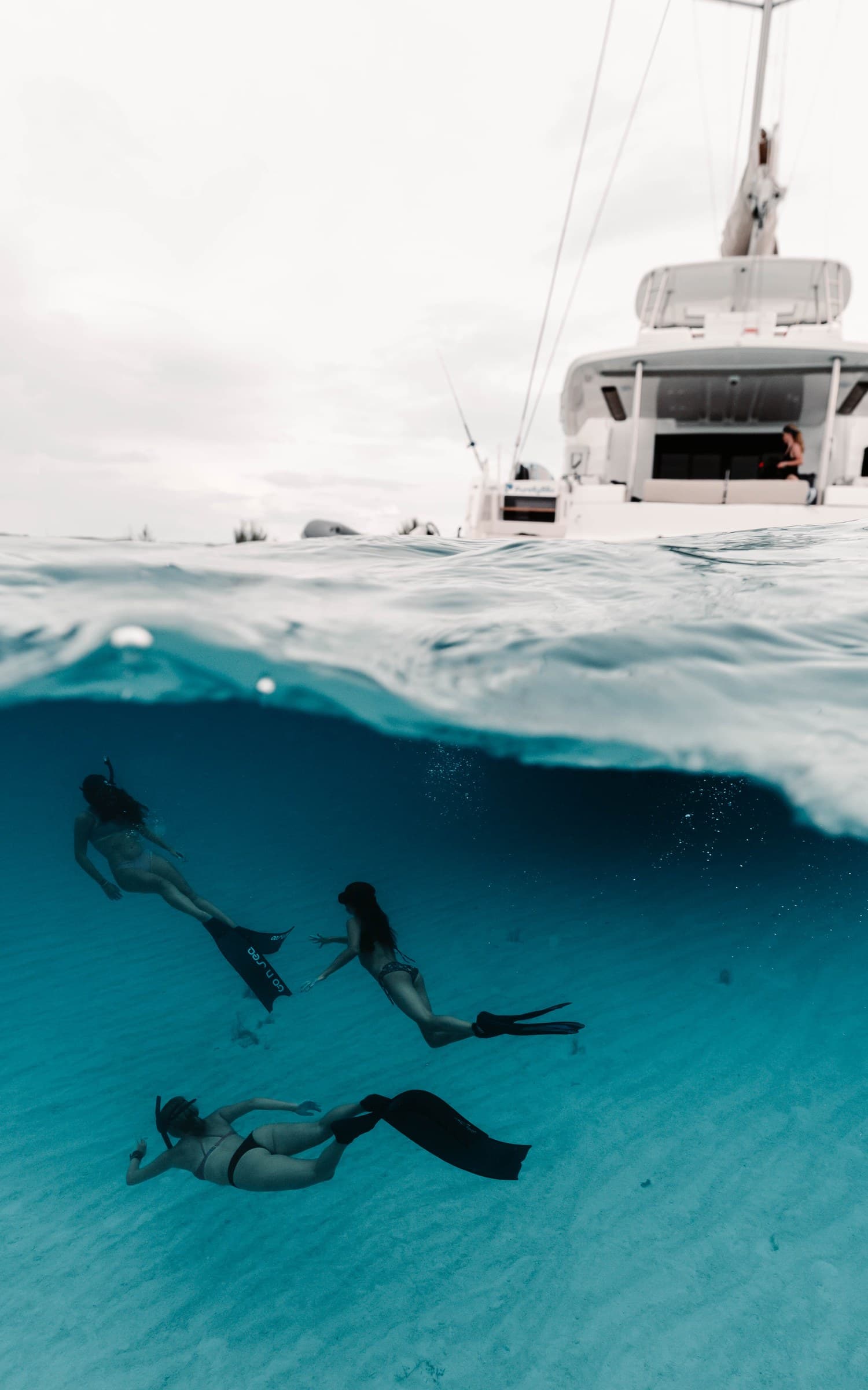 Split over-under: three freedivers beneath a catamaran — Exumas